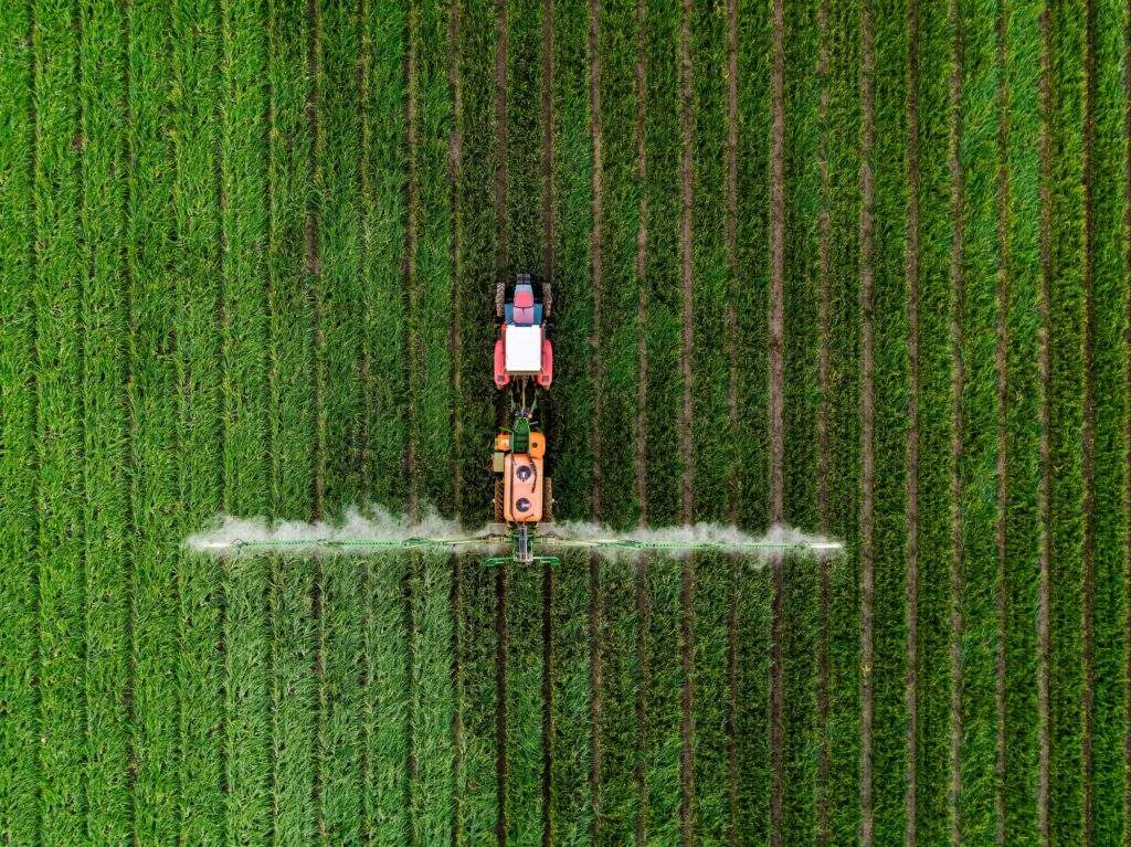 An aerial, top-down view of a red and orange tractor driving through a vibrant green field. The tractor is pulling a large spraying apparatus with long arms extending to both sides, emitting a fine white mist over the neat, vertical rows of crops. The symmetrical lines of the field create a textured, repetitive pattern across the entire frame.