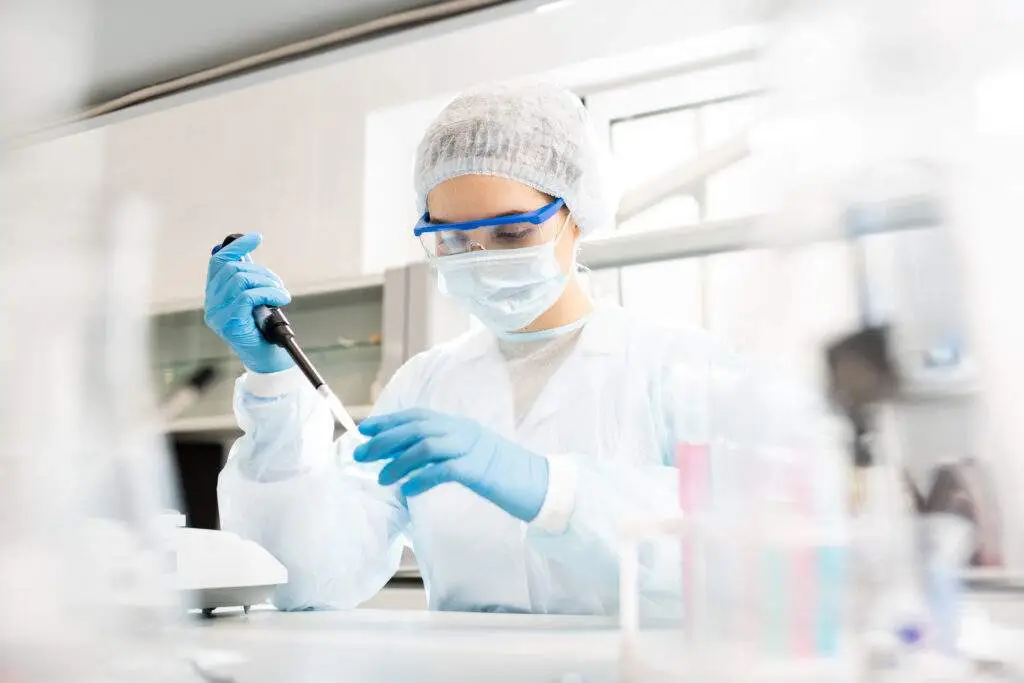 Serious concentrated female microbiologist in sterile clothing and safety goggles sitting at table and dropping reagent in petri dish while doing research in laboratory .