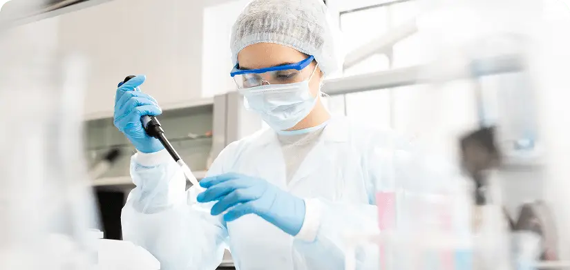 Serious concentrated female microbiologist in sterile clothing and safety goggles sitting at table and dropping reagent in petri dish while doing research in laboratory.