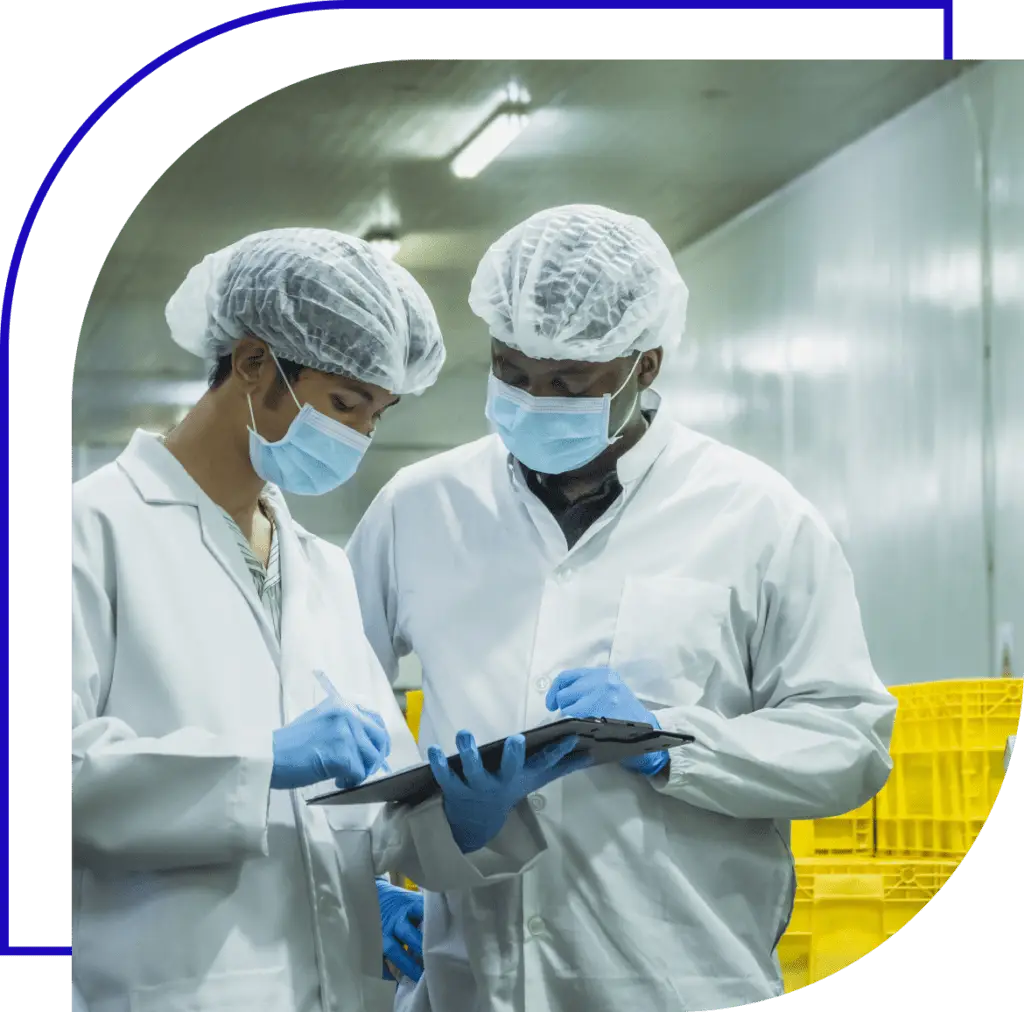 Two workers in a cleanroom or food processing facility wear white lab coats, hairnets, face masks, and blue gloves while inspecting a clipboard. One worker writes on the document while the other looks on attentively in an industrial setting with yellow crates in the background.