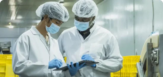 Two workers in a cleanroom or food processing facility wear white lab coats, hairnets, face masks, and blue gloves while inspecting a clipboard. One worker writes on the document while the other looks on attentively in an industrial setting with yellow crates in the background.