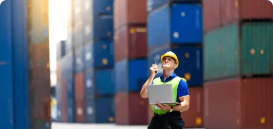 An outdoor shot of a logistics worker standing in a shipping container yard. The man is wearing a yellow hard hat, a bright green safety vest over a blue shirt, and dark work trousers. He is holding an open laptop in one hand and a walkie-talkie in the other, looking upward as if inspecting the towering stacks of multicolored (blue, red, and green) shipping containers that line the background. The scene is brightly lit by natural sunlight, creating a slight lens flare in the upper left corner.