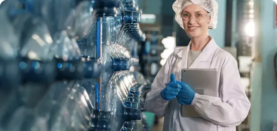 A technician in a white lab coat and hairnet stands in a water purification facility, holding a tablet and looking at the camera while surrounded by large water bottles and machinery.
