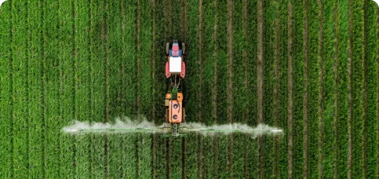 An aerial, top-down view of a red and orange tractor driving through a vibrant green field. The tractor is pulling a large spraying apparatus with long arms extending to both sides, emitting a fine white mist over the neat, vertical rows of crops. The symmetrical lines of the field create a textured, repetitive pattern across the entire frame.